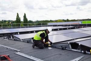 An engineer installing solar panels on the roof of the sports hall at Pediswell Leisure Centre, with Perdiswell Park visible in the distance.