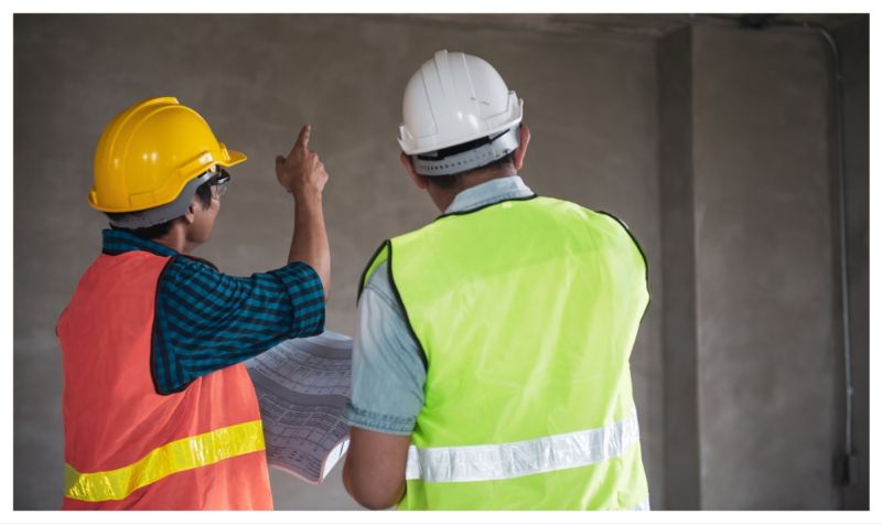 Two people in safety hats and hi-vis jackets with one pointing at a wall