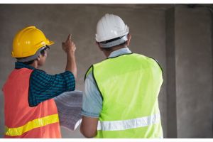 Two people in safety hats and hi-vis jackets with one pointing at a wall