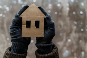 A pair of hands wearing thick gloves, holding a piece of cardboard in the shape of a house. Snow is falling in the background
