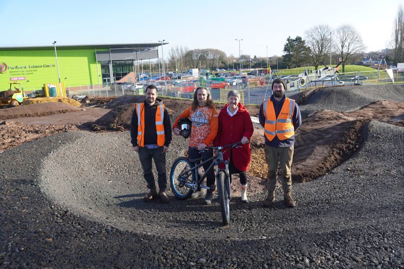 Four people, one holding a bike, standing in front of an unfinished pump track