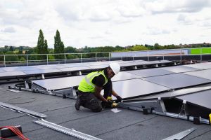 An engineer installing solar panels on the roof of the sports hall at Pediswell Leisure Centre, with Perdiswell Park visible in the distance.